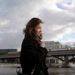 A young woman in a winter coat smiling while walking by a bridge in Berlin.